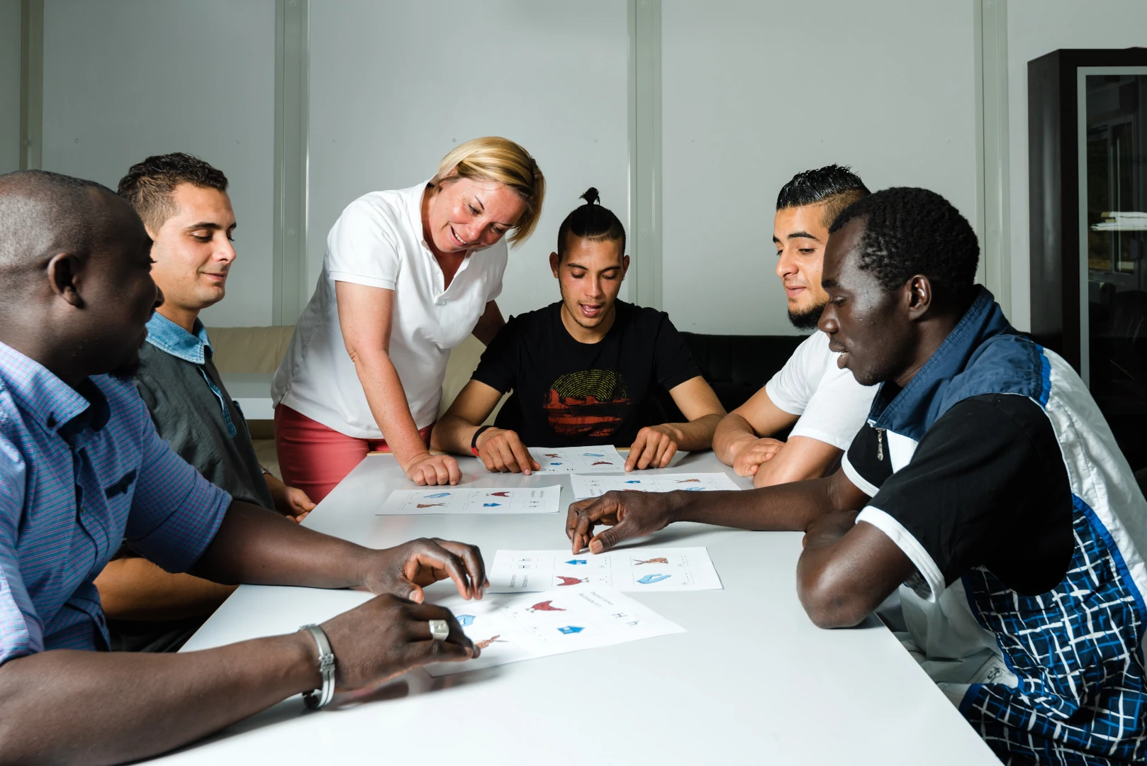 A group of men from different countries gathered around a table with a woman giving them a basic English lesson